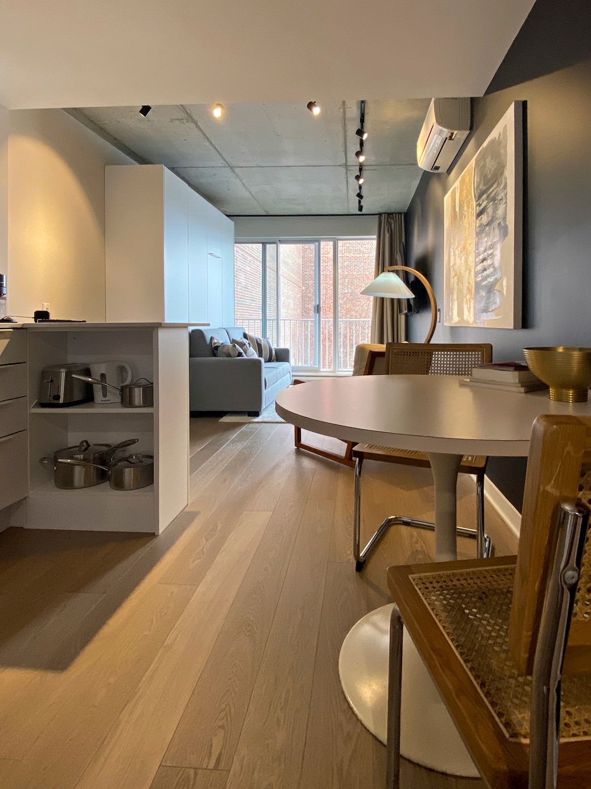 A modern living space features a round dining table accompanied by wooden chairs. A cozy sofa is visible in the background, complemented by large glass doors that allow natural light to fill the room. Subtle artwork and contemporary decor enhance the overall aesthetic.