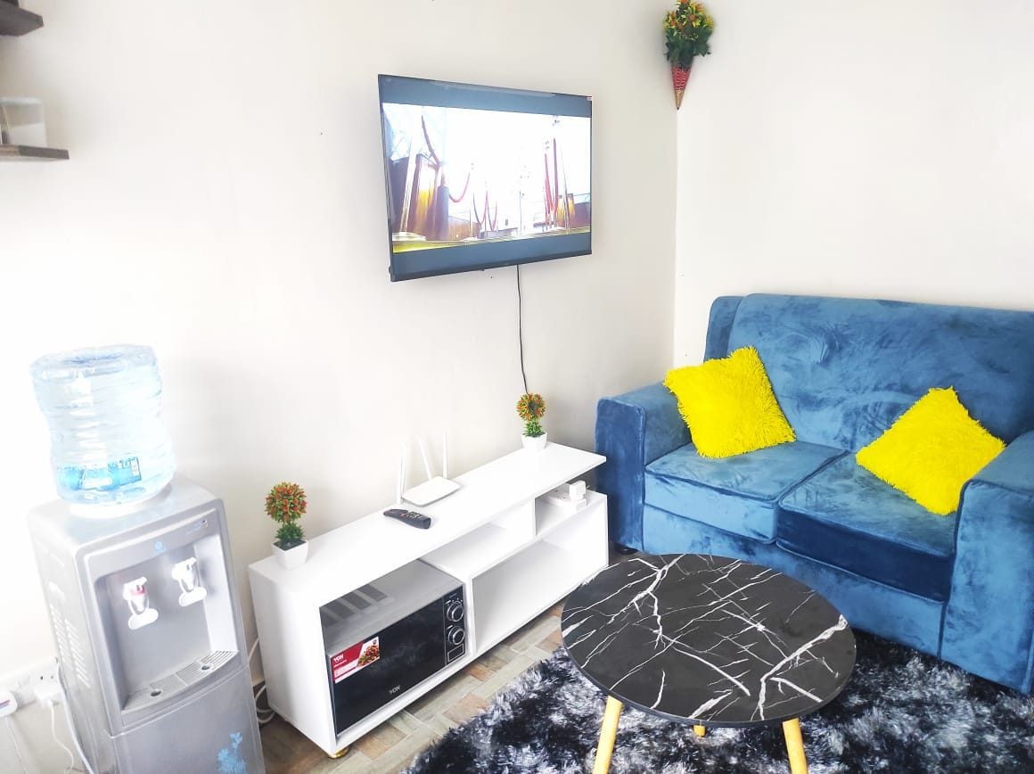 A cozy living area features a blue sofa adorned with yellow cushions. A dark marbled coffee table is positioned in front, while a television is mounted on the wall, providing entertainment options. A water dispenser and decorative plants enhance the inviting space.