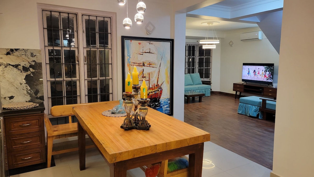 A spacious dining area features a large wooden table set for meals, surrounded by light-colored seating. Decorative candles are positioned at the center, while vibrant artwork depicting sailing ships adorns the wall. A view into the adjoining living area, with a TV and additional seating, is visible.