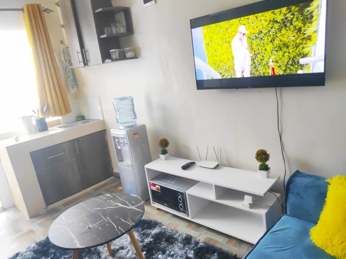 A modern living area is presented, featuring a comfortable blue sofa and a round marble-patterned table. A flat-screen TV is mounted on the wall, and a water dispenser stands next to a small kitchen space. Bright yellow curtains frame a window that allows natural light to enter.
