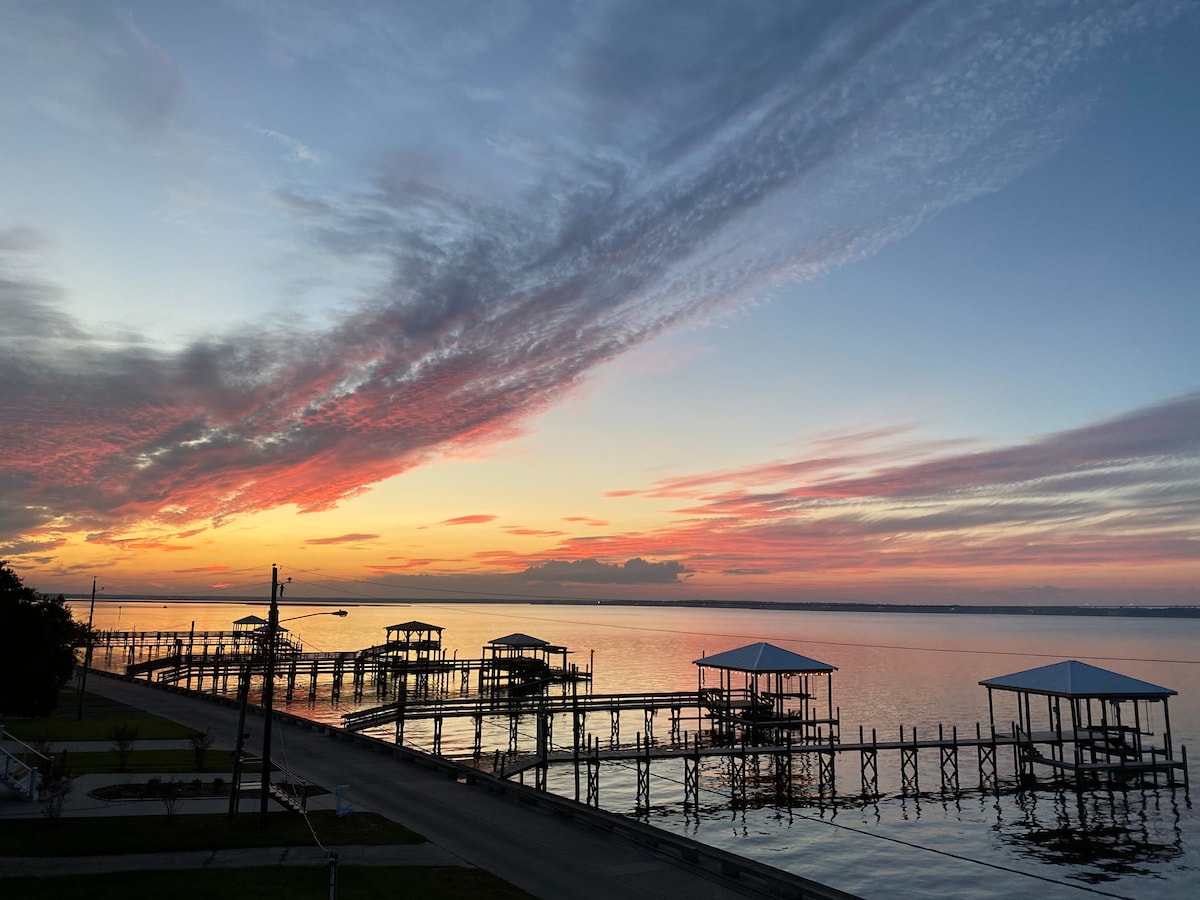 A serene waterfront view is captured at dusk, showcasing vibrant colors in the sky above calm waters. The scene features multiple piers extending into the bay, framed by gentle clouds and a well-maintained shoreline.