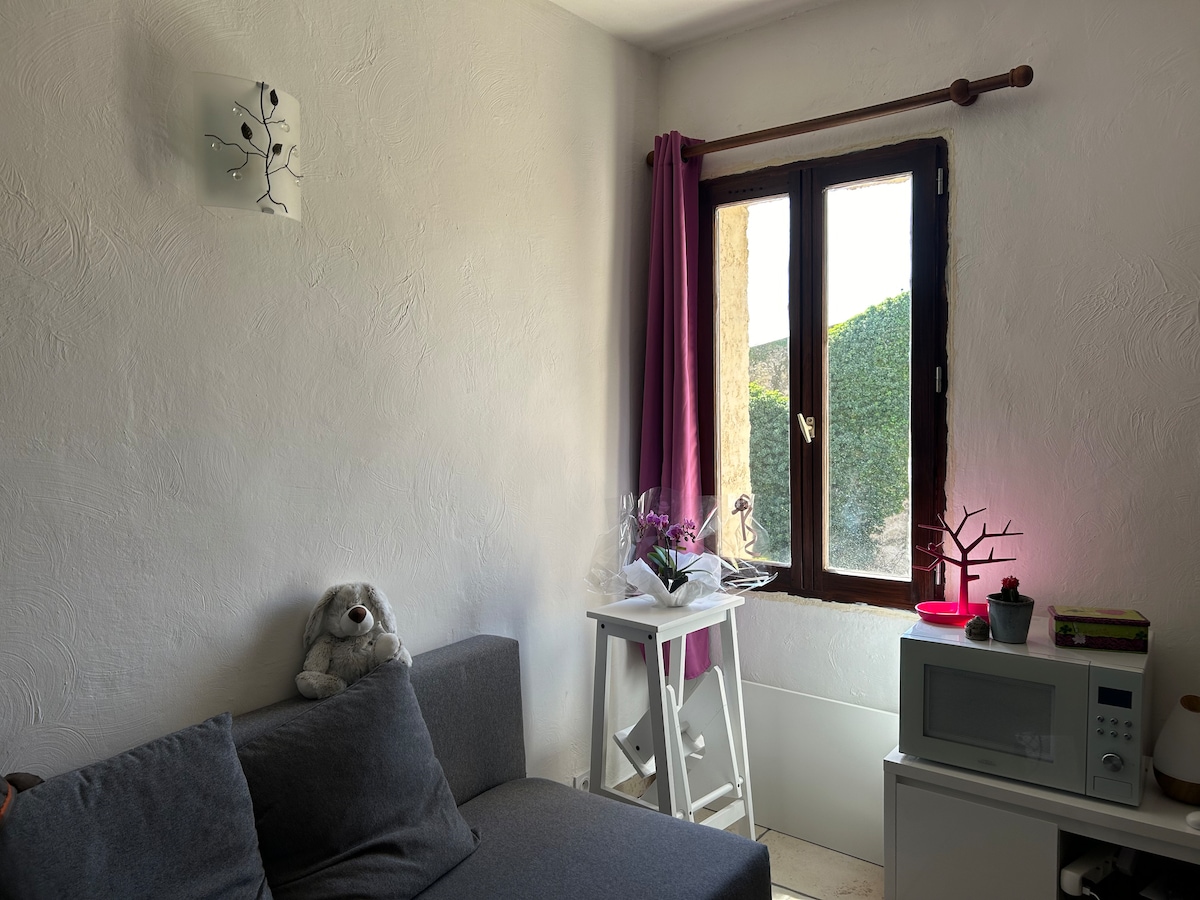 A cozy corner of the living area features a grey sofa and a white side table adorned with a small plant and decorative items. Sunlight filters through a window draped with sheer curtains, illuminating the space and highlighting a subtle wall decoration.