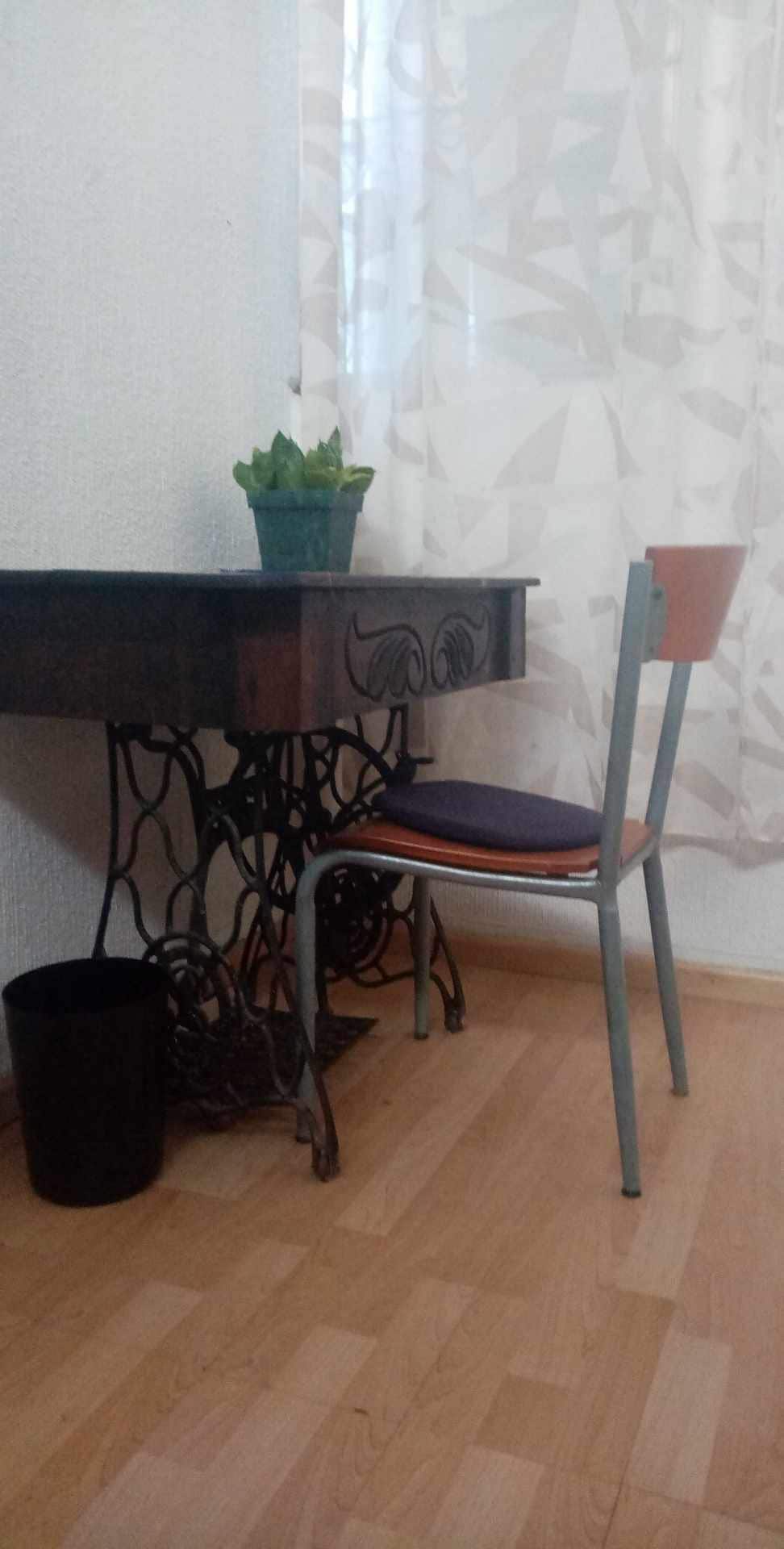 A compact workspace is presented with a decorative table featuring an intricate base. A chair with an orange seat is positioned nearby, accompanied by a small green plant on the table. Natural light filters through sheer curtains, creating a bright atmosphere.