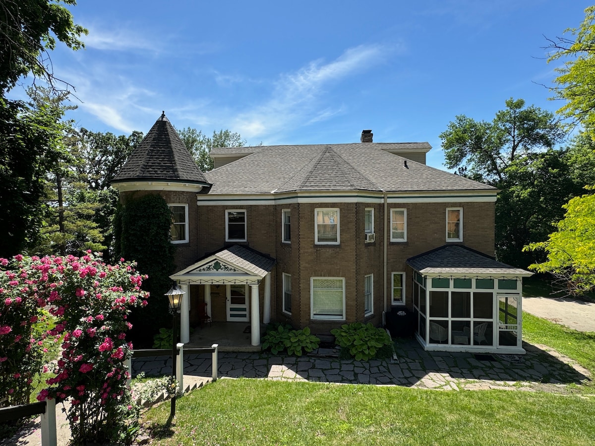 The exterior of the castle is captured under a clear blue sky. A stone pathway leads to the front entrance, framed by colorful flower bushes and well-maintained greenery. Unique architectural features include a turret and a covered porch, providing a welcoming entrance.