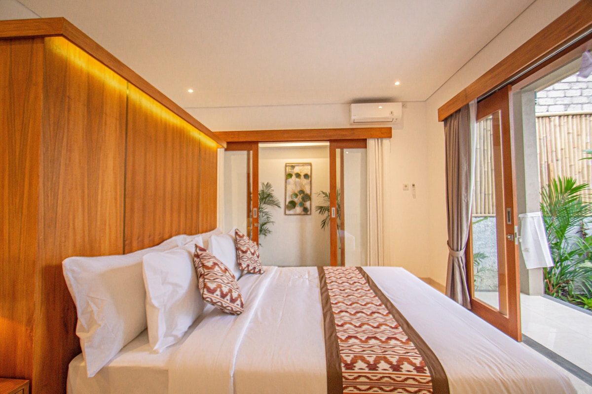 A bright bedroom features two spacious beds, dressed in crisp white linens and decorative throw pillows. Large sliding glass doors provide access to a private outdoor area, while warm wooden accents complement the overall design.