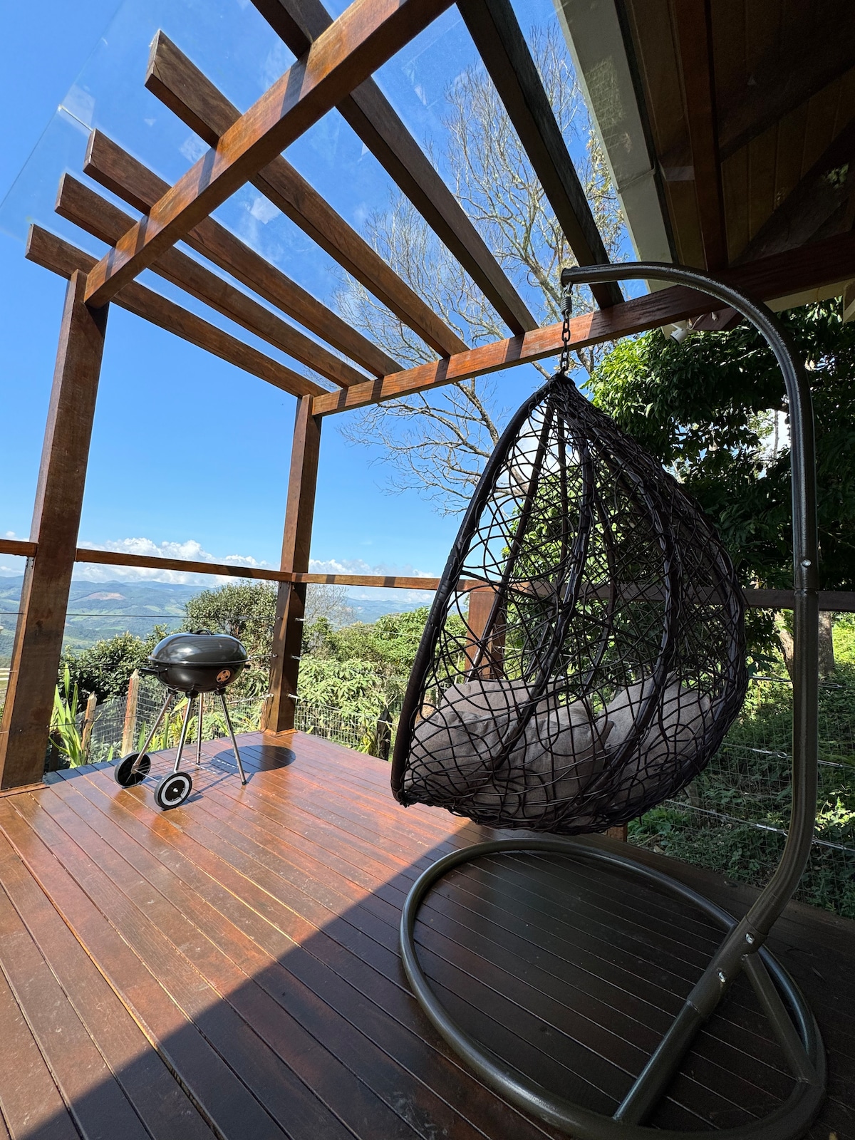 An outdoor seating area features a hanging chair with a cushioned interior, offering expansive views of the surrounding landscape. A charcoal grill is positioned nearby, and the wooden deck is bordered by a lattice structure with a transparent roof, allowing sunlight to filter through.
