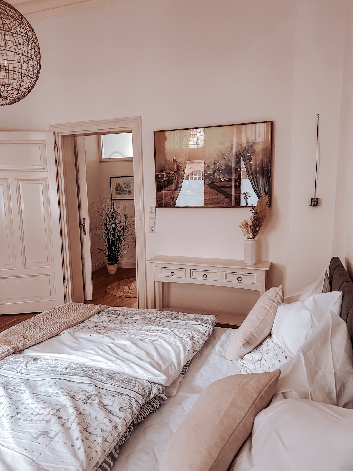 A softly lit bedroom features a spacious bed adorned with decorative pillows and a textured blanket. A neutral-toned dresser sits against the wall, complemented by wall art and a minimalistic vase. Natural light filters through a nearby window, enhancing the room's serene atmosphere.