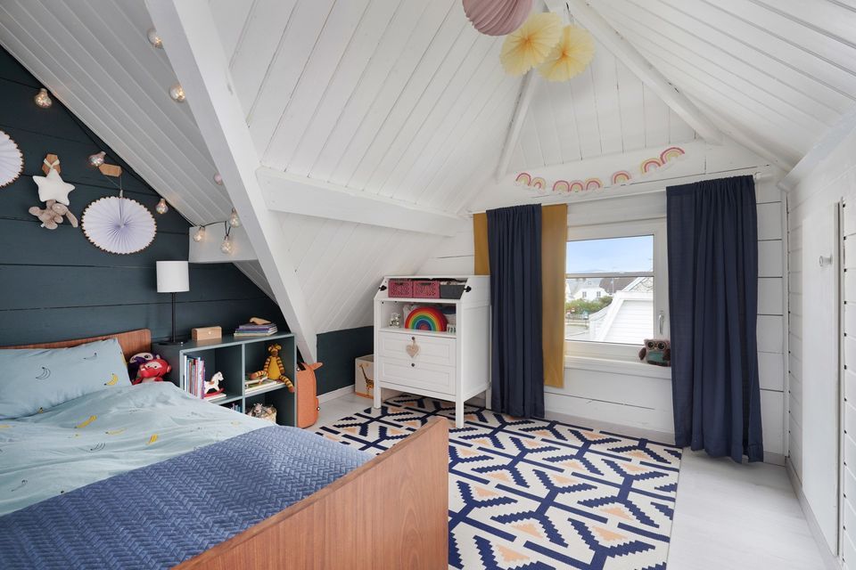 A room with sloped ceilings features a large bed with light bedding. A colorful area rug covers the floor, while a bright window is framed by dark curtains. Decorative wall elements, including paper fans and a playful storage unit, add character to the space.