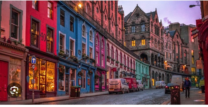 A charming street is lined with colorful buildings painted in shades of blue, pink, and green. The buildings showcase a variety of windows and a richly textured facade. Street lamps illuminate the cobbled road, enhancing the inviting atmosphere of this unique Edinburgh neighborhood.