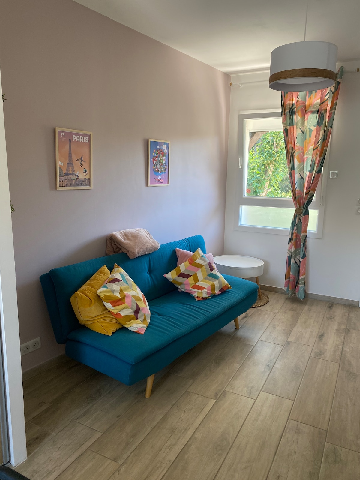 A cozy seating area features a teal sofa adorned with colorful cushions. A small round table sits beside it, and a window allows natural light to fill the space. Light curtains with a floral pattern frame the view of the greenery outside.