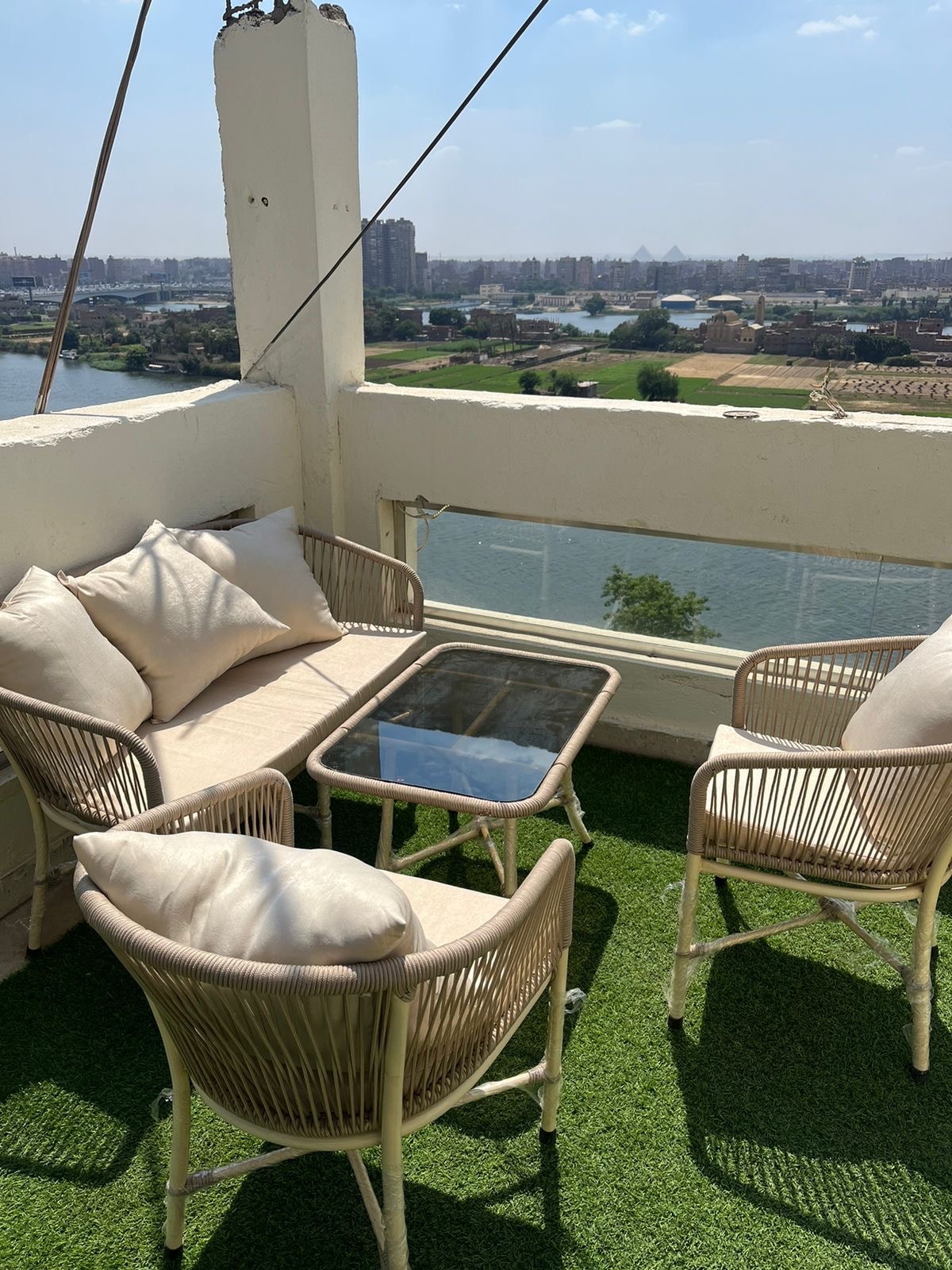 A rooftop seating area features a glass table surrounded by four woven chairs, each adorned with light cushions. A view of the Nile River and distant pyramids is visible, framed by the balcony's white walls and green artificial grass flooring.