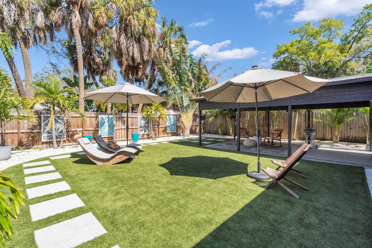 The backyard features a landscaped area with artificial grass, two shaded lounge chairs, and large umbrellas. A pathway of stepping stones leads across the space, flanked by tropical plants and palm trees, creating a relaxing outdoor environment.
