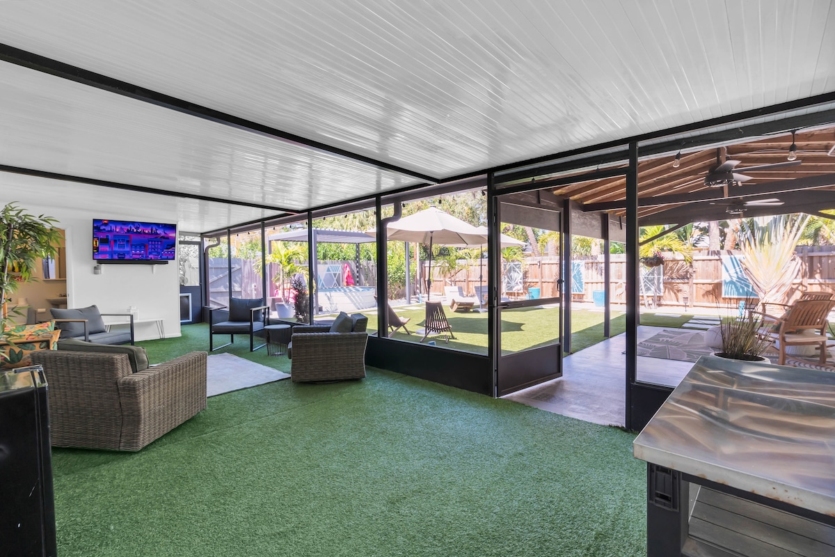 A spacious screened-in patio features synthetic grass flooring, providing a comfortable area for relaxation. Seating arrangements include a couple of lounge chairs and a sofa, creating an inviting space. A flat-screen TV is mounted on the wall for entertainment, with views extending to the outdoor pool area.