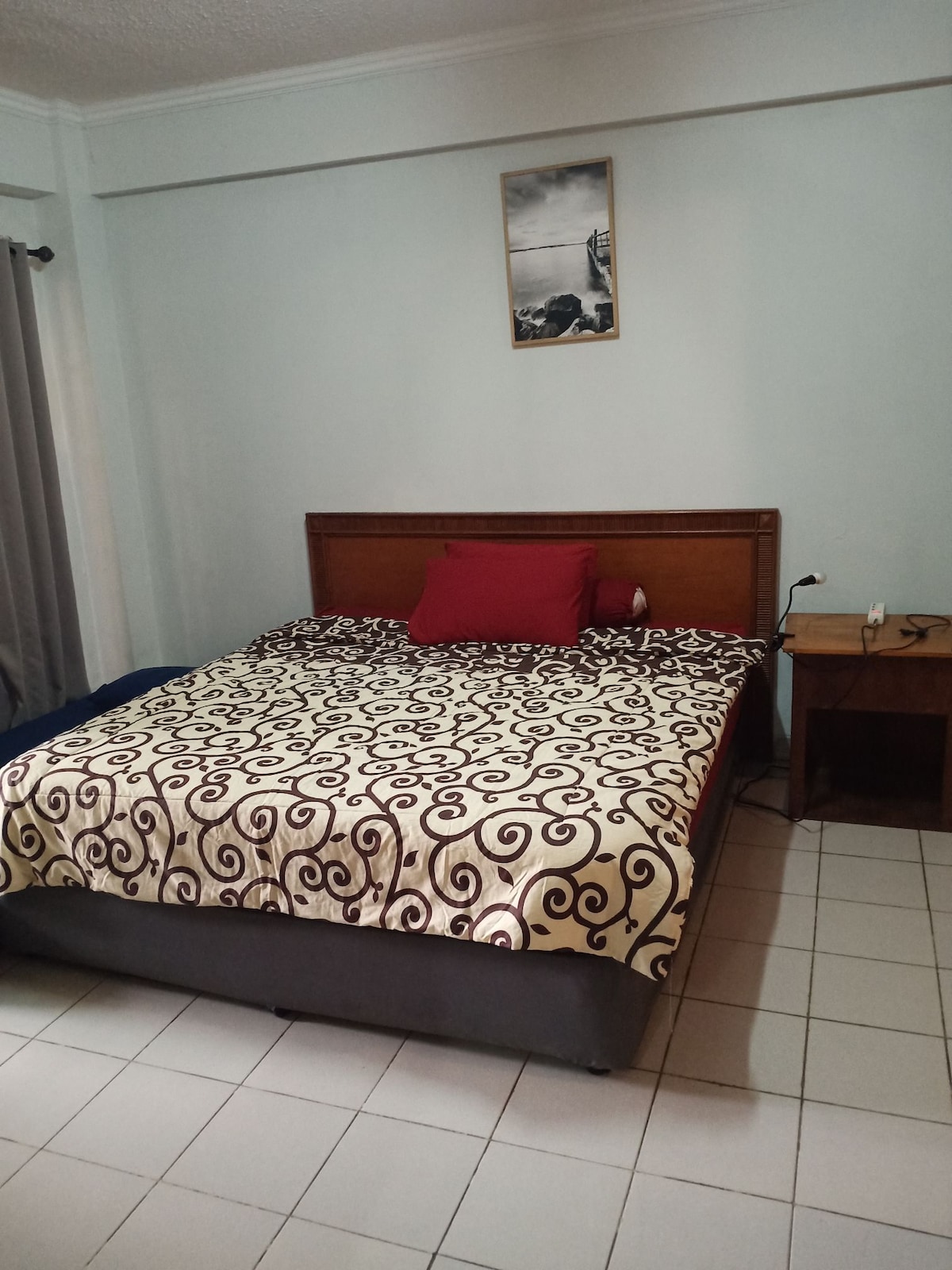 An inviting sleeping area features a spacious bed adorned with a patterned blanket and a single decorative pillow. Nearby, a wooden nightstand holds a lamp and a charging station. Soft grey curtains frame a window, allowing natural light to illuminate the room.