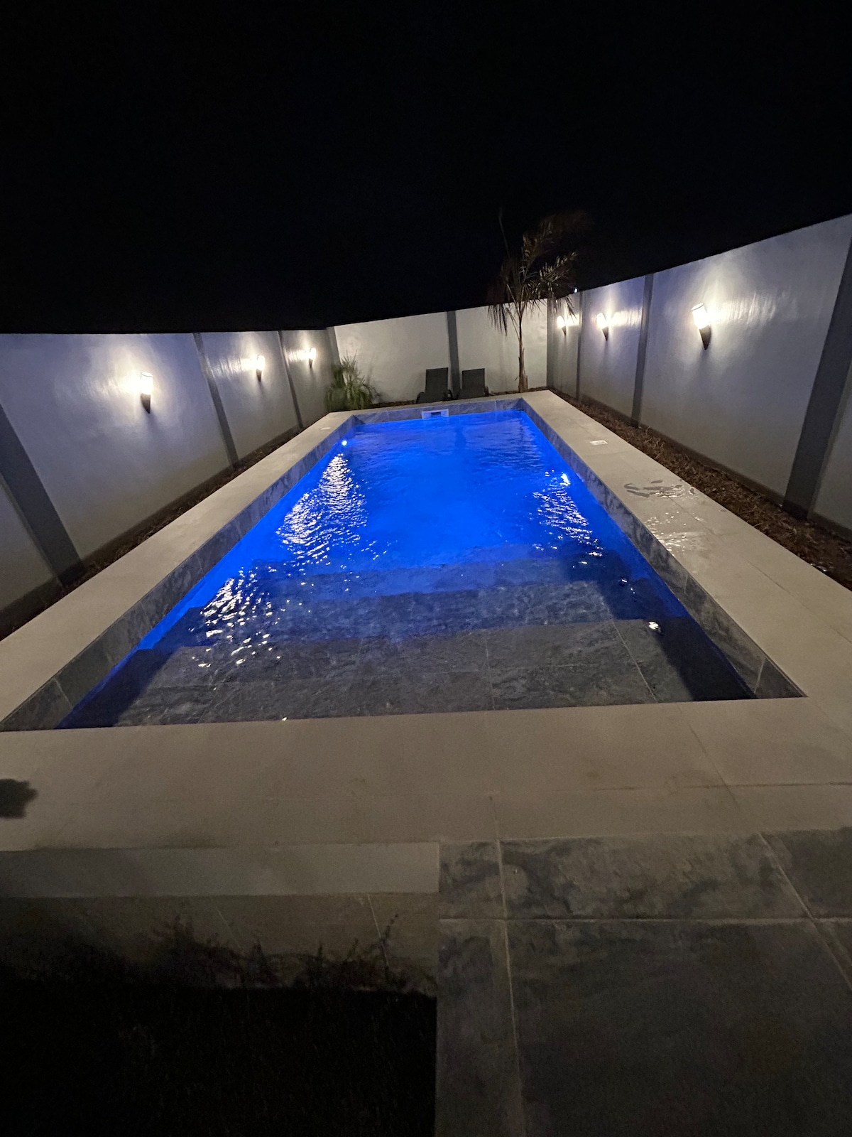 A modern swimming pool is illuminated in shades of blue, set within a serene outdoor space. Soft lighting along the walls enhances the tranquil atmosphere, while the clean lines of the surrounding area provide a peaceful escape for relaxation.