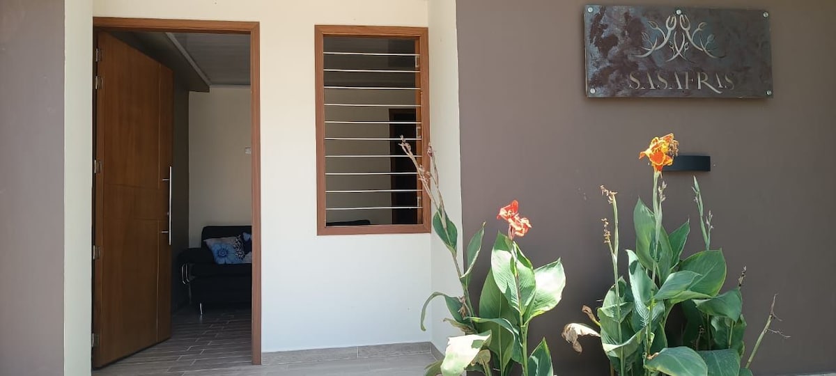The image depicts the entrance of a modern apartment, featuring wooden doors and a window with metal slats. Vibrant green plants with orange flowers are positioned in the foreground, enhancing the welcoming appearance of the entrance area.