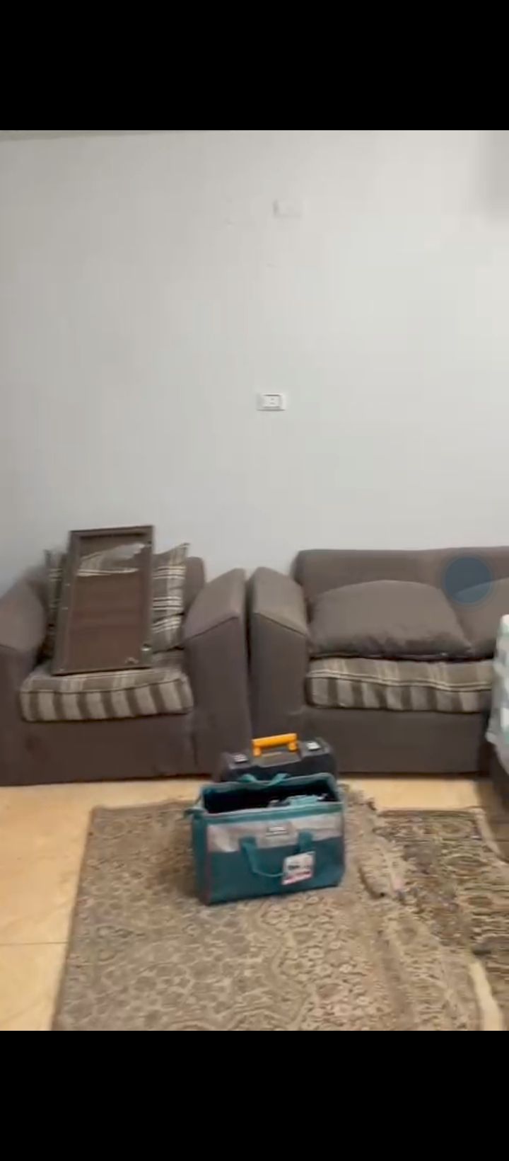 Two comfortable armchairs with striped cushions are positioned next to a light-colored bed. A utility bag rests on a patterned area rug, while a framed piece leans against the wall nearby. A neutral wall complements the decor.