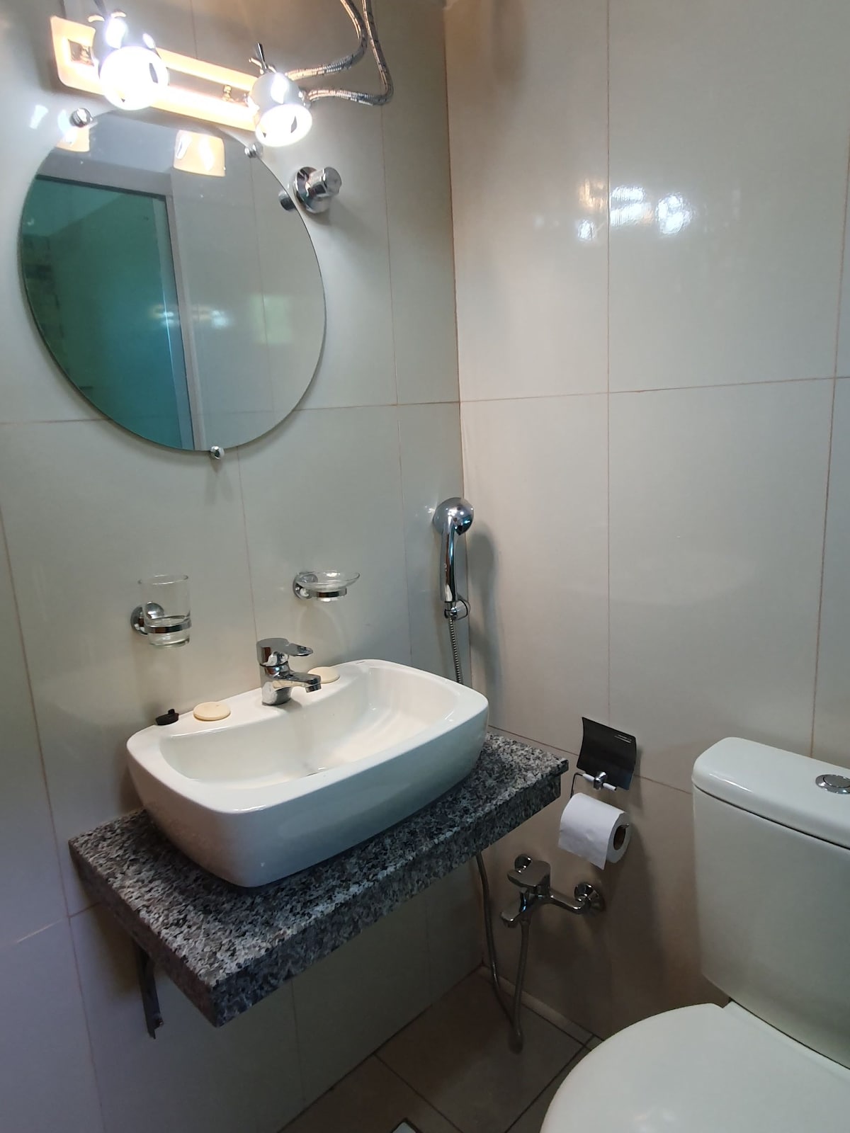 A modern bathroom is shown, featuring a white sink positioned above a granite countertop. A circular mirror is mounted on the wall above the sink. Toilet fixtures are visible, along with a roll of toilet paper on a holder. Natural light is softly filtered into the space.
