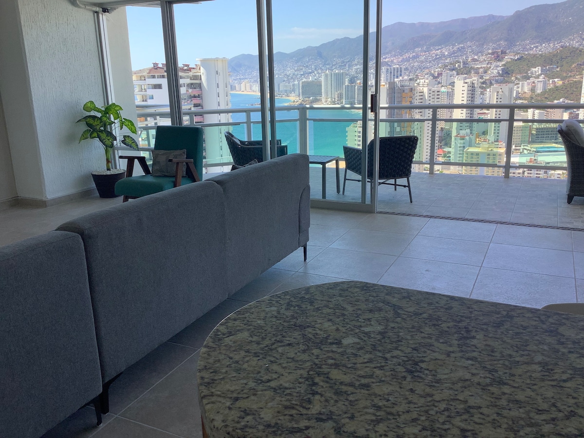 A spacious living area features a large gray sofa facing a floor-to-ceiling glass wall, providing stunning views of the bay. A round dining table with stone surface is included, while seating arrangements with dark wicker chairs and greenery can be seen in the background.