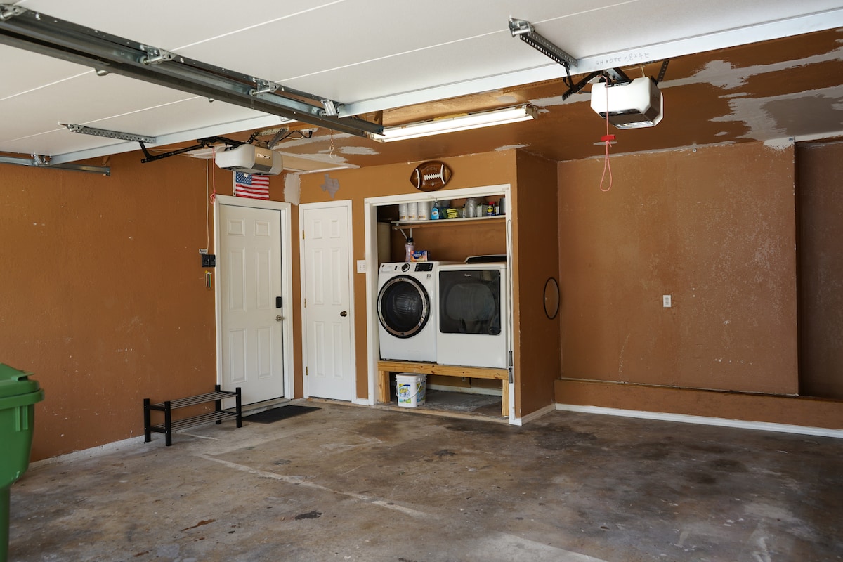 A garage space features a beige color scheme with concrete flooring and minimal clutter. A washing machine and dryer are located against one wall, while a door leads to the exterior. An American flag hangs from the ceiling, adding a touch of character.