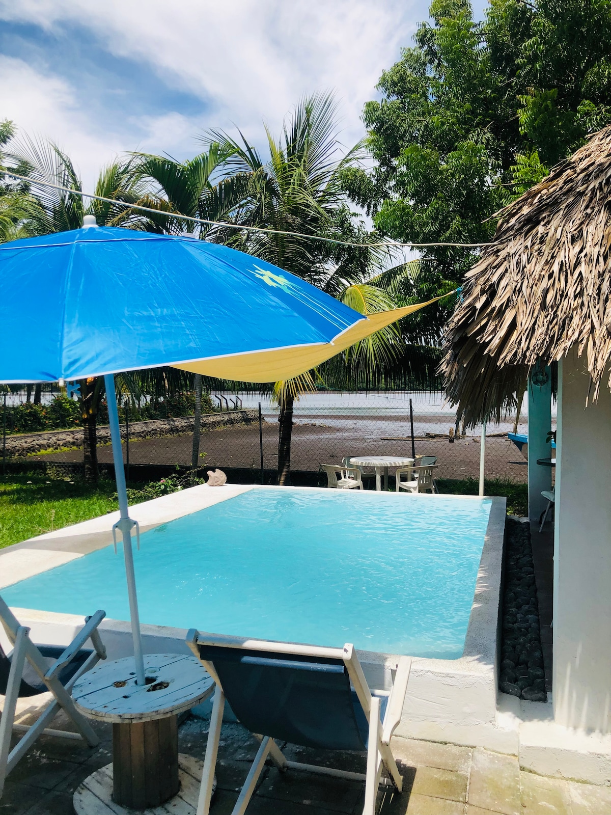 A private swimming pool is positioned next to a thatched-roof structure, surrounded by lush greenery and palm trees. A large blue umbrella provides shade over a few outdoor chairs, while the serene estuary is visible in the background.