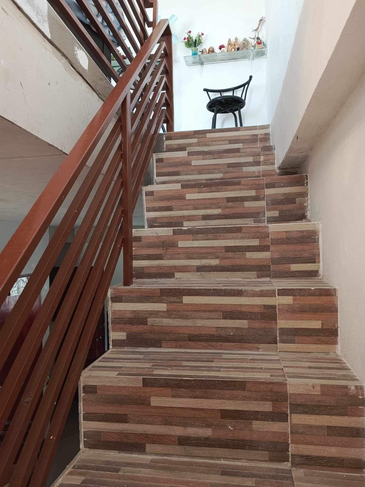 A set of stairs is shown, featuring a patterned wood-like tile design. A sturdy wooden railing runs alongside the steps. At the top of the stairs, a single black chair is positioned against the wall, which highlights the modern aesthetic of the space.