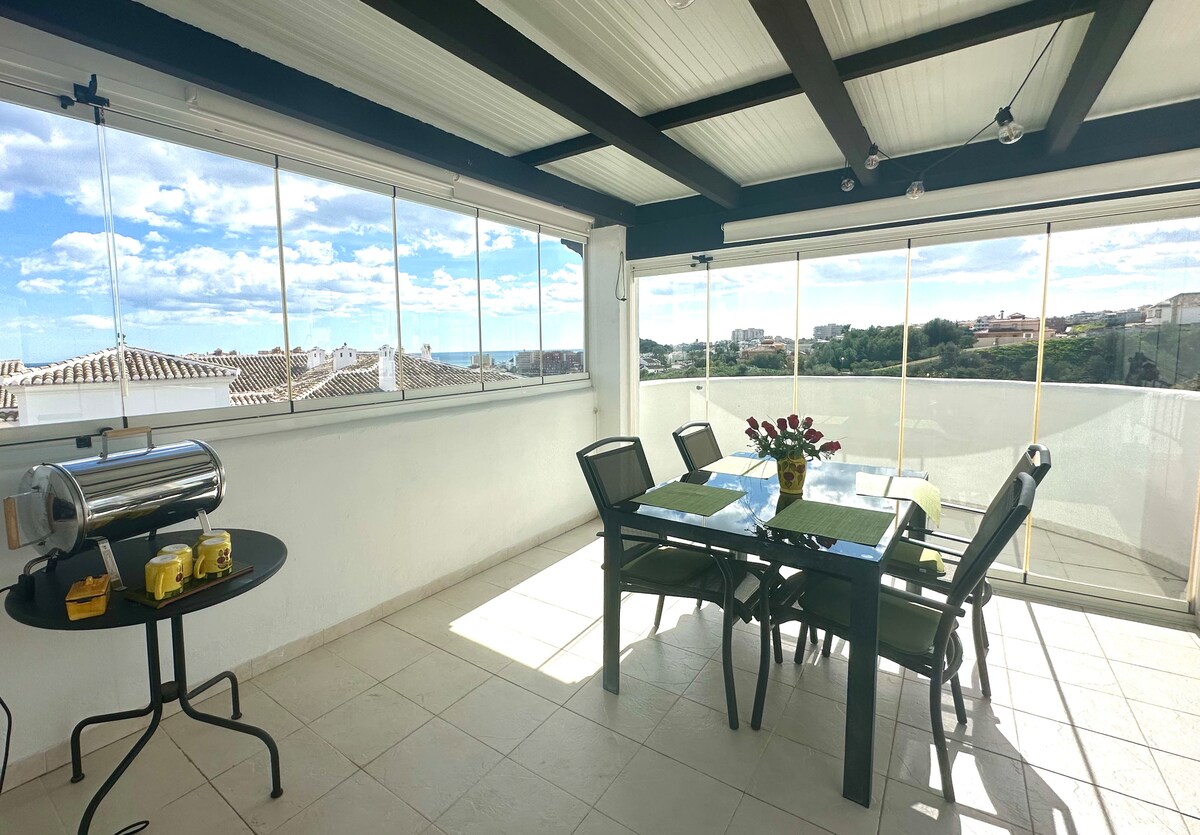 A bright terrace area features a dining table with chairs and a decorative vase of roses. A barbecue grill is positioned on the side. Large glass panels provide panoramic views of the surrounding landscape, allowing natural light to fill the space.