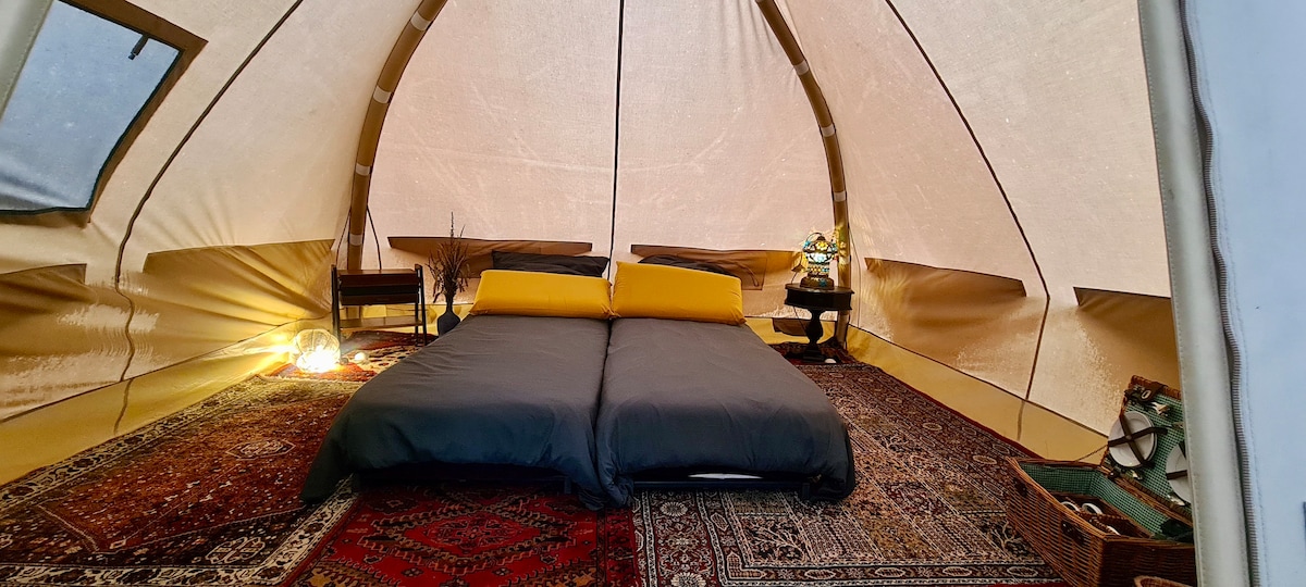 A spacious glamping tent features a queen size bed dressed with fresh linens. Warm lighting highlights the interior, complemented by decorative rugs. Wooden side tables are positioned next to the bed, creating a cozy atmosphere within the canvas walls.