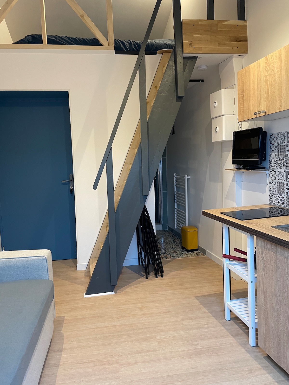 A spiral staircase leads to a lofted sleeping area, adorned with a cozy mattress. The kitchenette features modern cabinetry and utensils. A towel radiator is visible along the wall, providing added comfort. Natural light illuminates the open space, enhancing the functional layout.