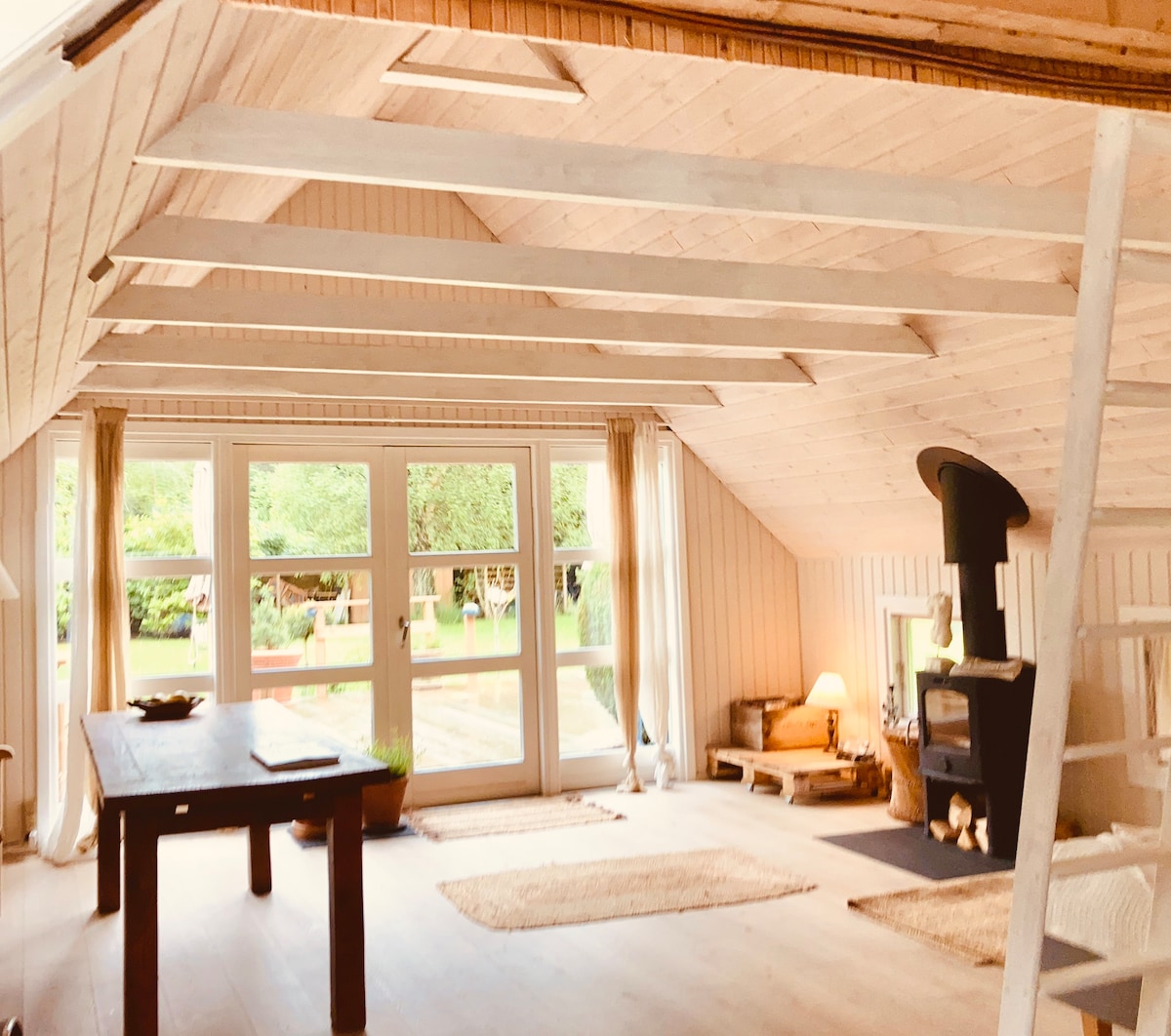 A light-filled living area features a high wooden ceiling with exposed beams. Large windows provide views of the garden, while a cozy fireplace and a wooden table offer inviting spots for relaxation. Soft area rugs enhance the warm aesthetic.