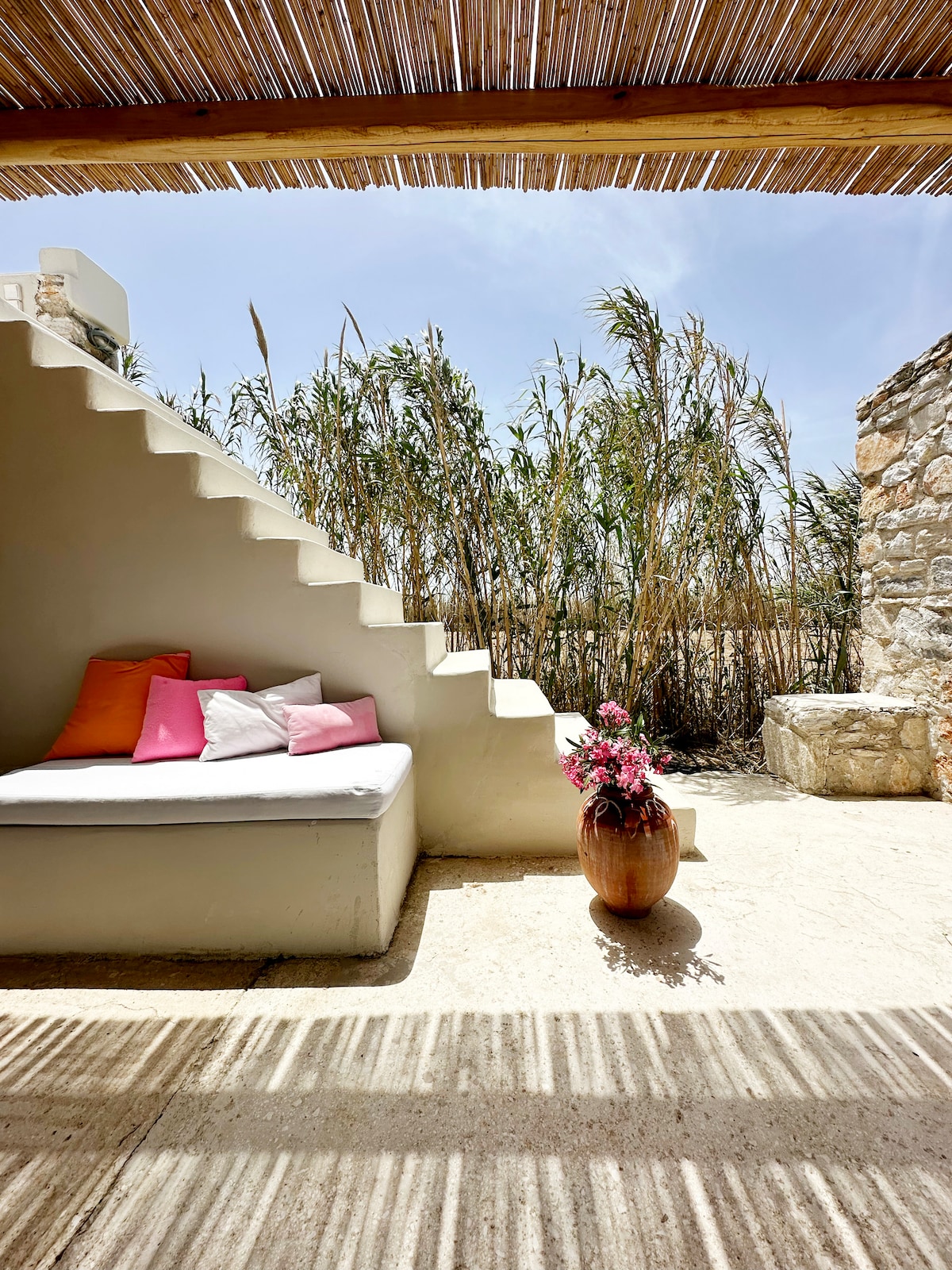A secluded outdoor seating area features a built-in bench adorned with colorful cushions. A decorative vase filled with flowers adds a touch of nature, while a stone wall provides a rustic backdrop. Sunlight casts patterned shadows across the space, enhancing the tranquil environment.