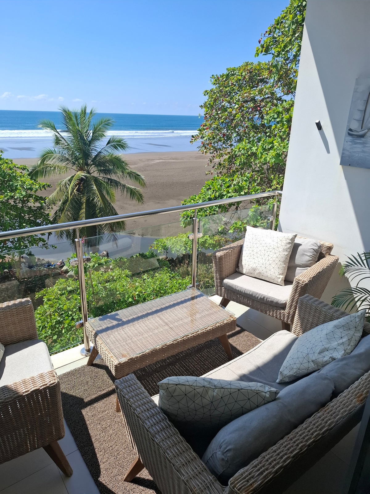 A covered balcony features comfortable outdoor seating, including two armchairs with cushions and a coffee table. Lush greenery is visible alongside the tranquil beach view and ocean waves. The clear blue sky complements the inviting atmosphere, creating a serene outdoor space.
