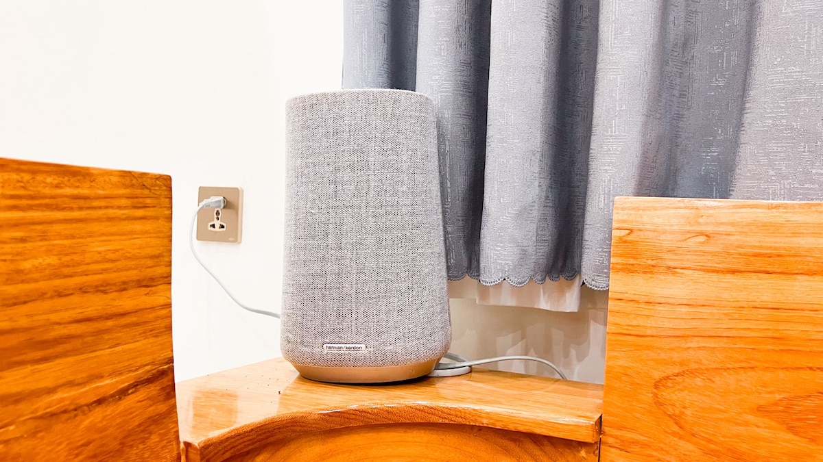 A modern speaker is positioned on a wooden side table, its grey fabric exterior complementing the warm tones of the wood. A grey curtain softly drapes in the background, adding a subtle texture to the setting.