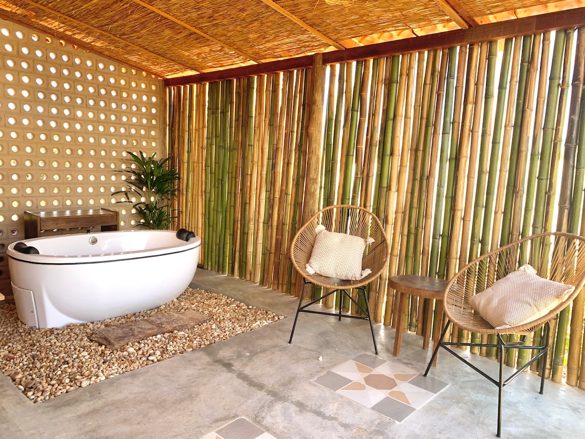 An inviting outdoor space features a freestanding round bathtub surrounded by pebbles and a bamboo wall for privacy. Two stylish chairs with cushions provide a comfortable seating area, enhancing the serene ambiance of the setting.