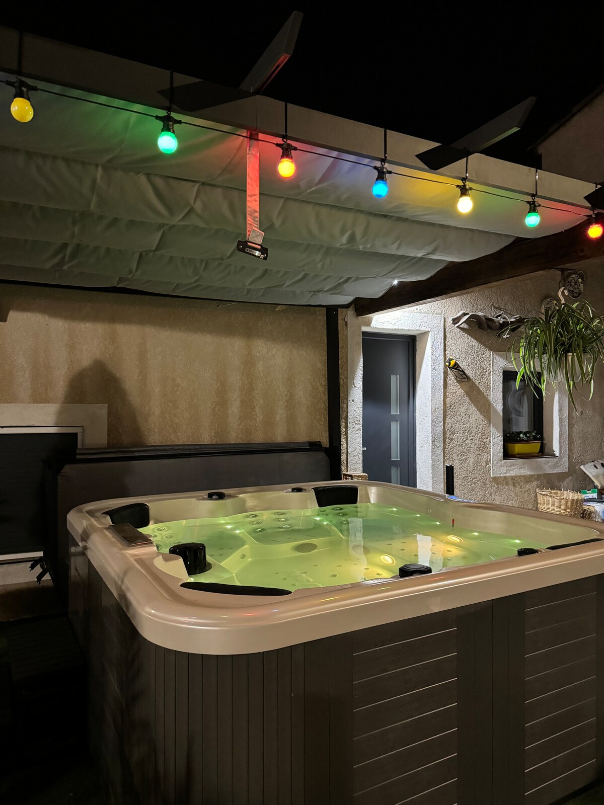 A spacious hot tub is positioned under a covered area, surrounded by colorful string lights that add subtle illumination. The setting is tranquil, inviting relaxation, with a wall featuring decorative elements and a door leading to an indoor space.