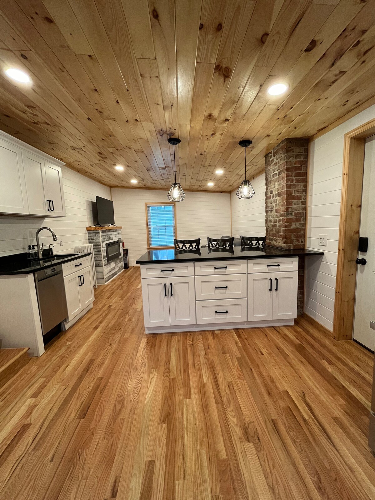 A newly renovated kitchen features a spacious layout with wooden flooring and a ceiling adorned with wooden panels. White cabinetry and a contrasting black countertop create an elegant contrast. Pendant lighting hangs above a central island, while a window allows natural light to fill the room.