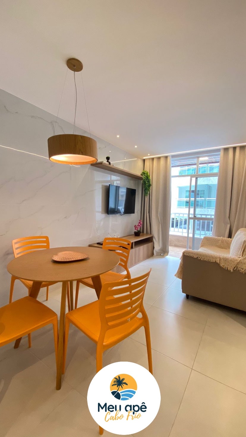 A dining area featuring a round table with four bright orange chairs is seen alongside a cozy seating space. Large windows provide natural light, and a sleek television unit is positioned on the wall. Neutral tones are complemented by subtle greenery.