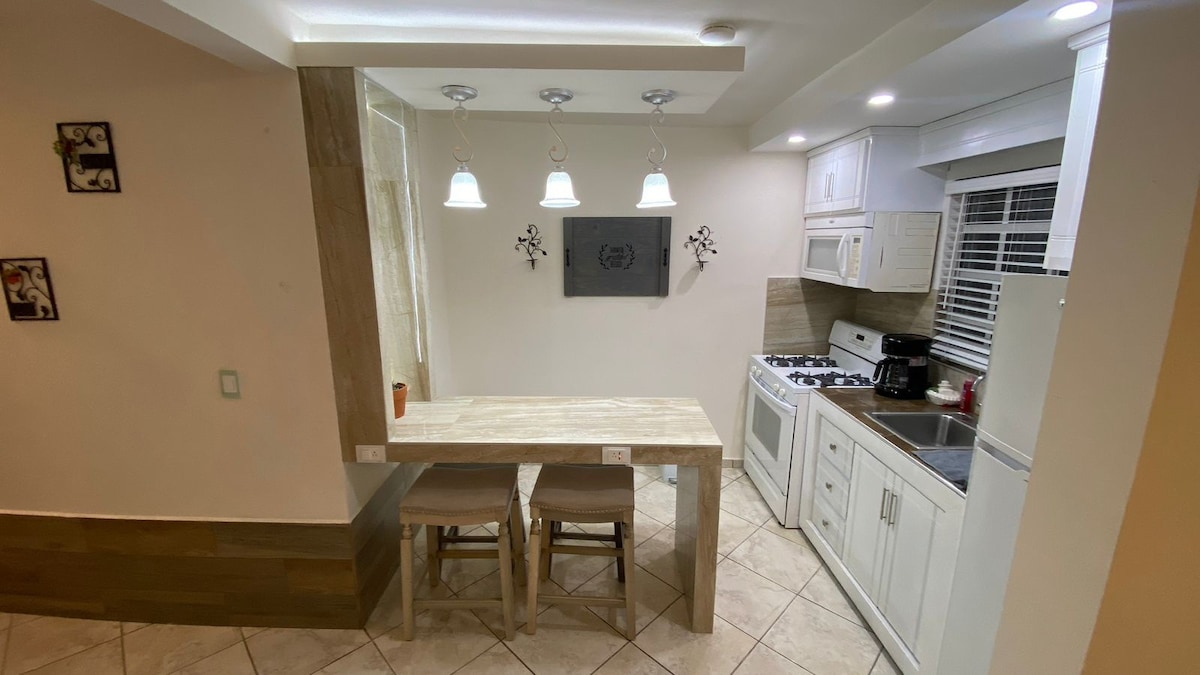 A modern kitchen area features a light-colored table with two stools and overhead pendant lighting. Sleek white cabinetry, a stainless-steel sink, and a gas stove are visible, complemented by tile flooring that enhances the space's functionality.