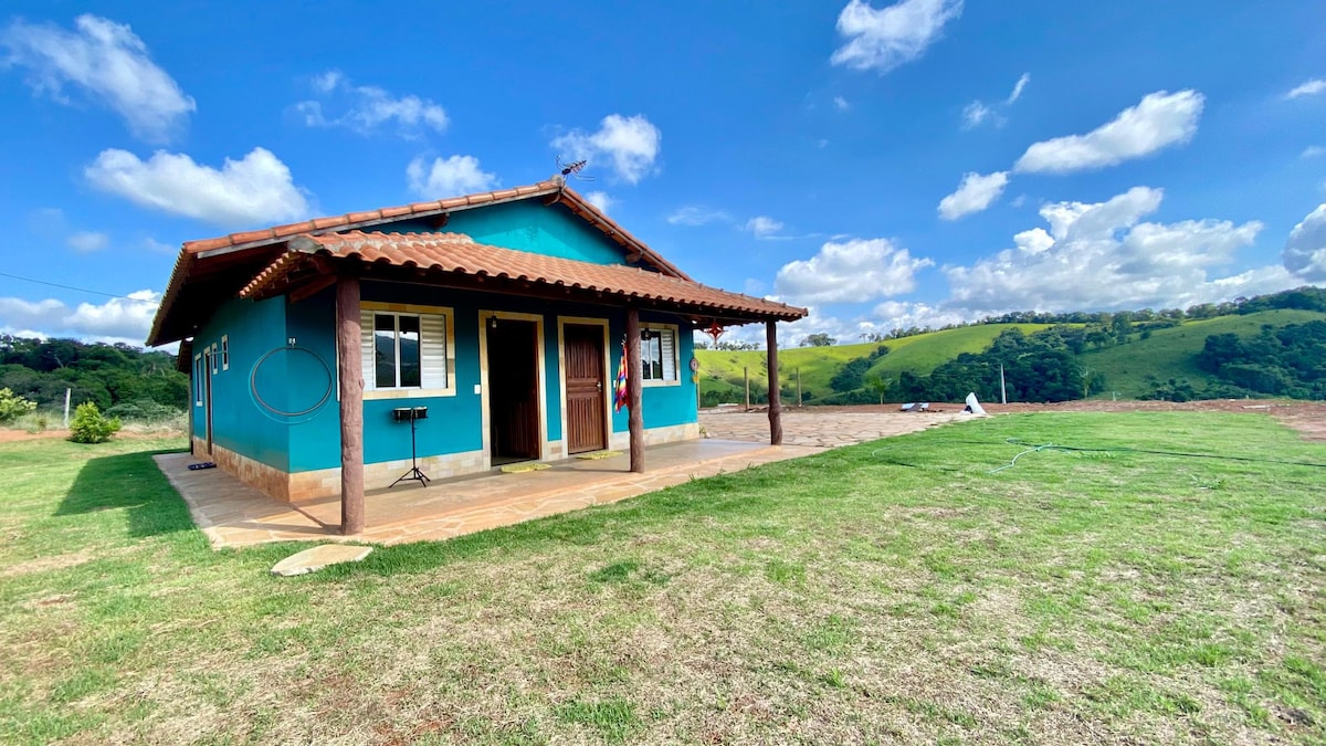 A brightly painted house with a blue exterior and brown roof is viewed from the grassy yard. The covered porch features two entrances, and a barbecue grill is visible on the side. Surrounding hills are seen in the background under a partly cloudy sky.