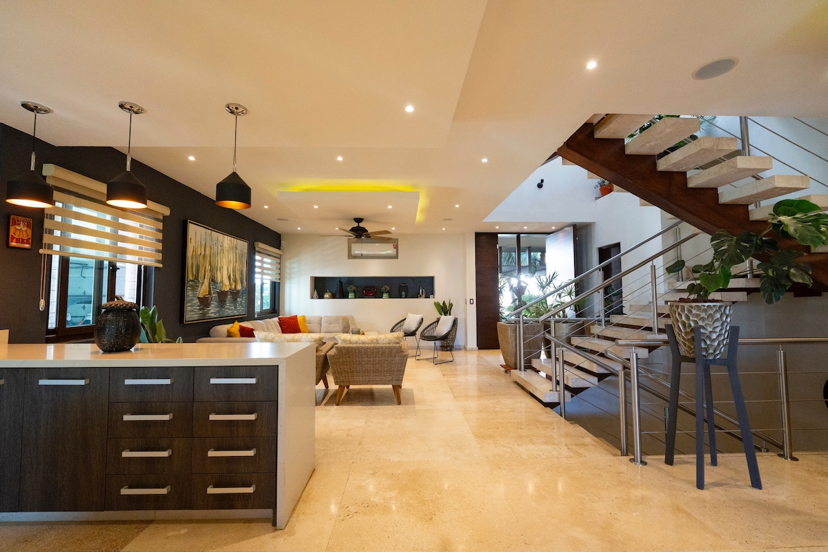 A spacious living area showcases a comfortable seating arrangement and modern architectural design. Large windows allow natural light, while contemporary lighting fixtures provide a warm ambiance. A sleek kitchen with a central island is visible, blending seamlessly with the dining space and staircase leading to upper levels.