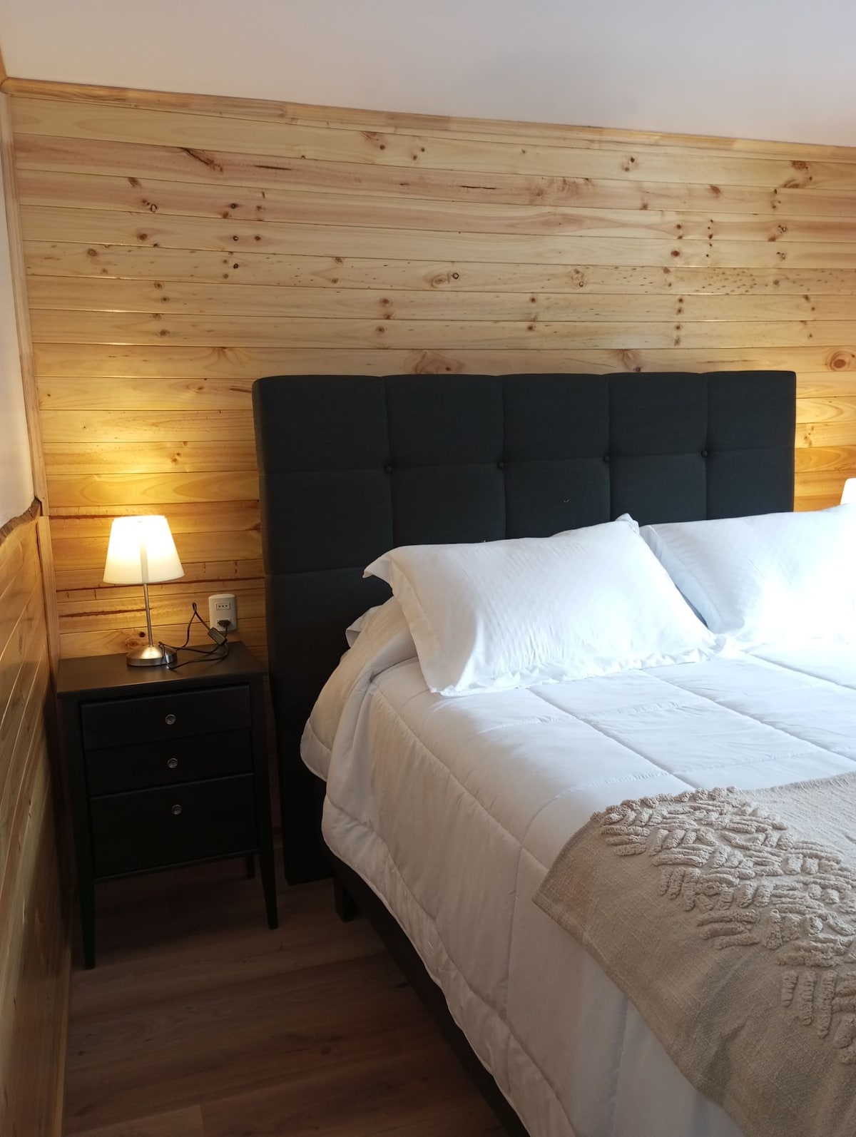 A cozy bedroom is featured with a dark upholstered headboard and a spacious bed dressed in crisp white linens. A bedside table with a lamp is positioned next to the bed, complementing the natural wood-paneled walls and creating a peaceful atmosphere.