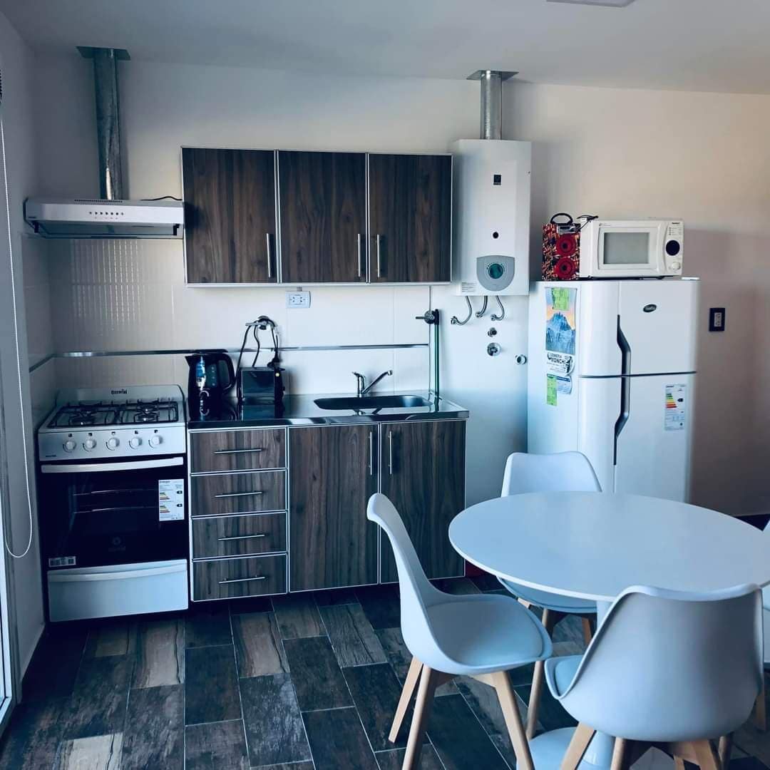 A modern kitchen area features wooden cabinets and stainless-steel appliances. A compact dining table with white chairs is positioned in front. The kitchen includes a gas stove, oven, refrigerator, and sink, with additional storage units and a microwave visible on the countertop.