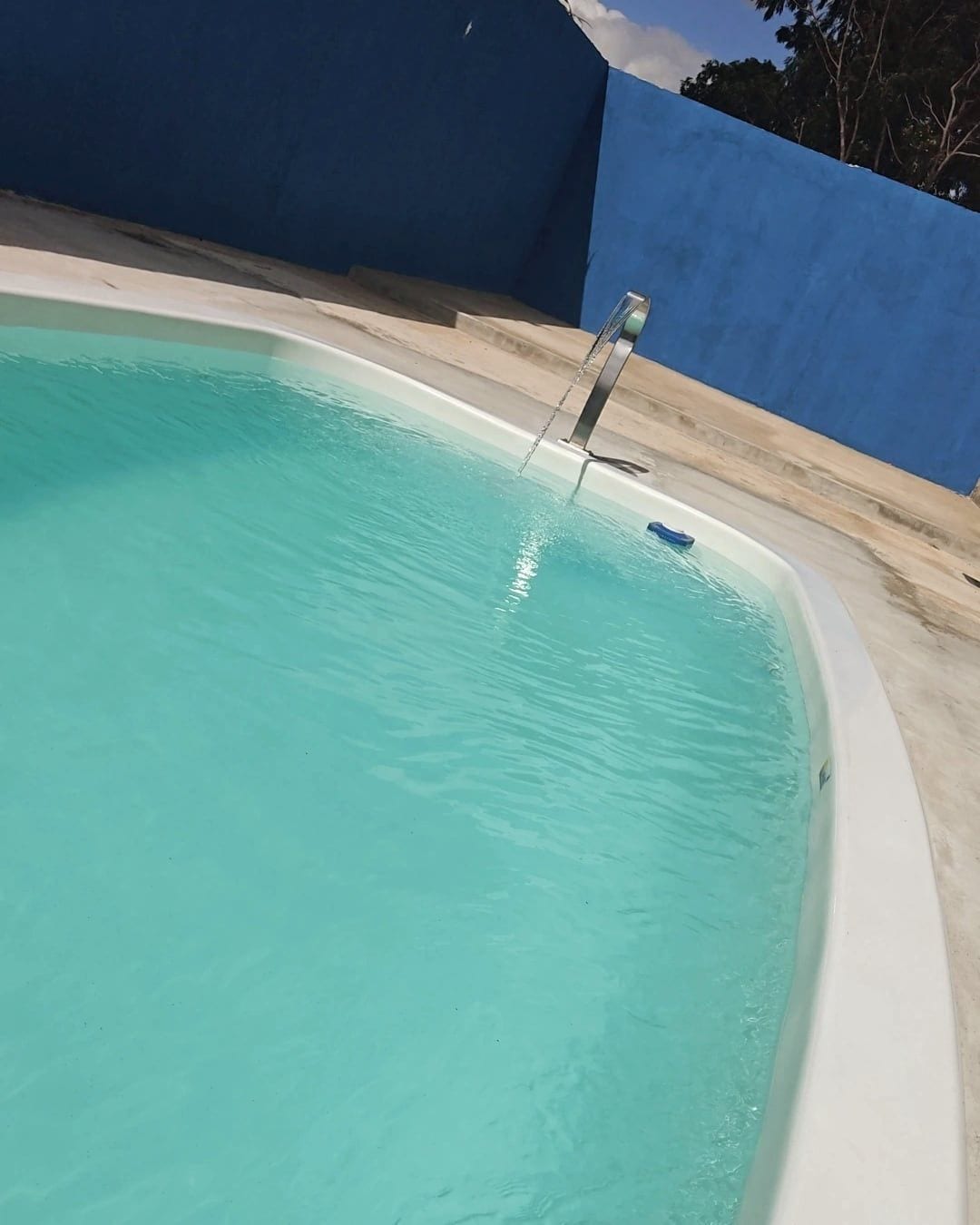 A refreshing swimming pool features clear turquoise water and a sleek curved shape. A gentle water feature is visible, adding a subtle dynamic to the serene setting. The surrounding area includes a smooth concrete deck against a bold blue wall, contributing to a vibrant ambiance.