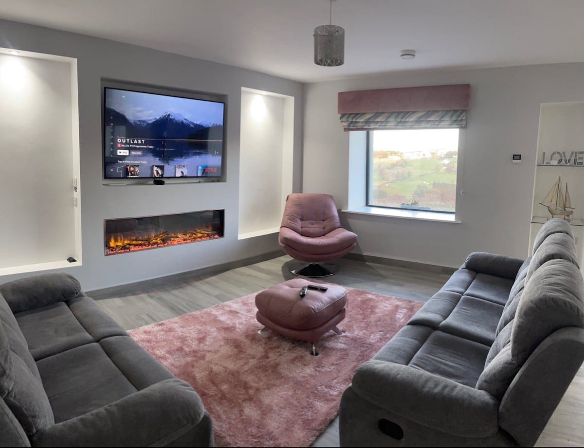A modern living area features a large wall-mounted television above a sleek electric fireplace. Two grey sofas face each other across a plush area rug. A pink accent chair adds a pop of color, while a window lets in natural light, framed by soft fabric blinds.