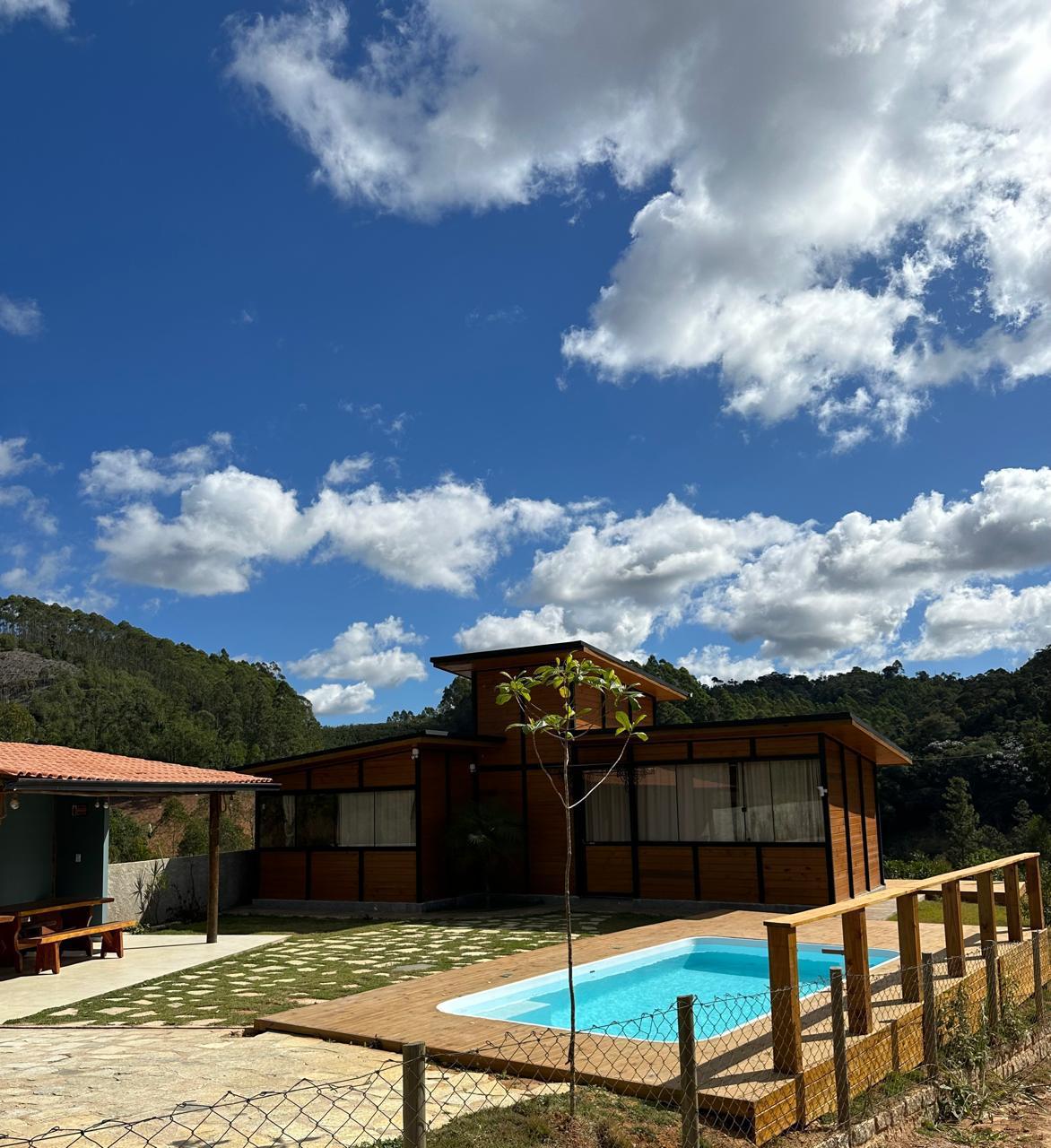 A wooden house is featured with large windows, allowing natural light to fill the interior. A small turquoise pool is set in front, surrounded by a grassy area. The backdrop of lush trees and a bright blue sky with clouds enhances the tranquil setting.