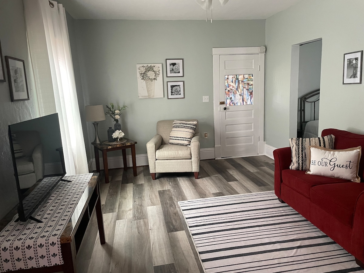 A well-lit living area features a comfortable red sofa and a cream armchair. A wooden table stands beside a lamp, while a flat-screen television is positioned on a low console. Framed artwork adorns the walls, and light-colored curtains gently drape over a window.