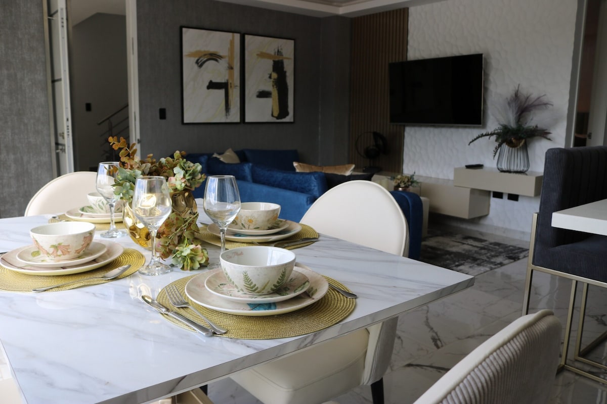 A dining table is set with elegant dinnerware, featuring decorative plates and glassware, accompanied by a decorative centerpiece. In the background, a cozy living area is visible, showcasing a blue sofa and artwork on the walls. Natural light illuminates the space.