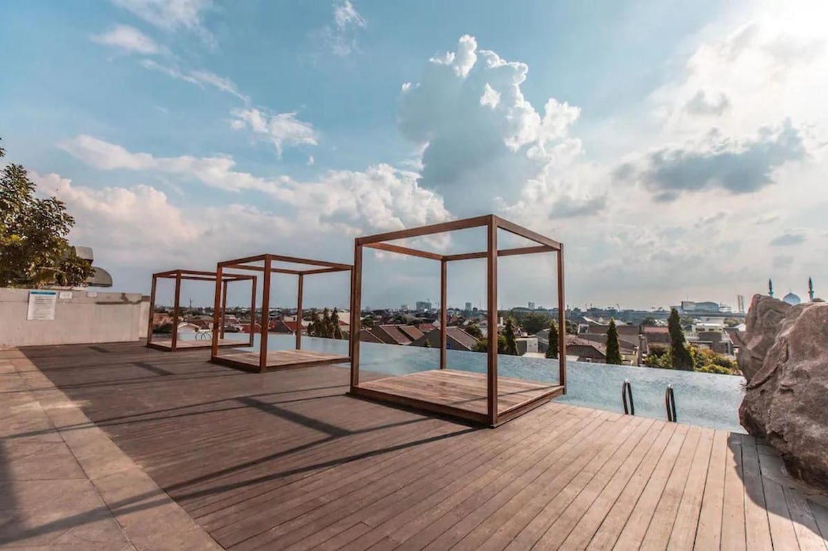 An open rooftop area features wooden platforms framed by sleek structures, offering panoramic views of the surrounding cityscape. A tranquil pool is visible at the edge, reflecting the cloudy sky above, while ample wooden decking creates a spacious setting for relaxation.