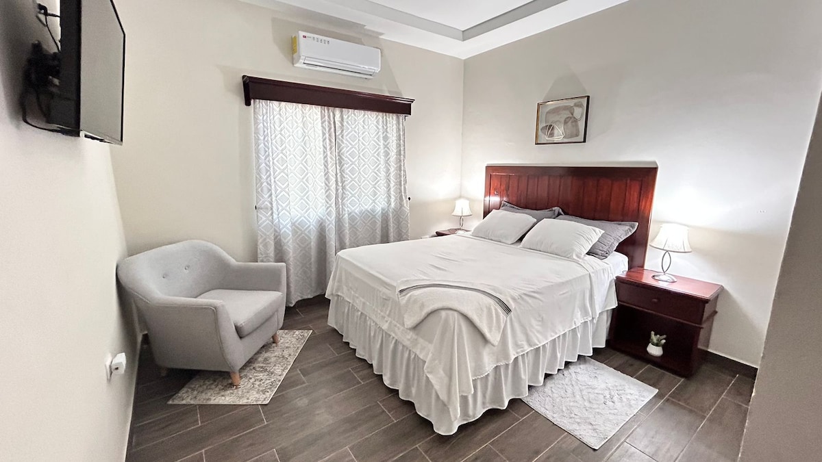 A comfortable sleeping area is highlighted by a spacious bed with clean white linens. An armchair sits in the corner beside a window, softly filtered by light curtains. Two bedside lamps provide gentle illumination, and a simple artwork adorns the wall above the bed.