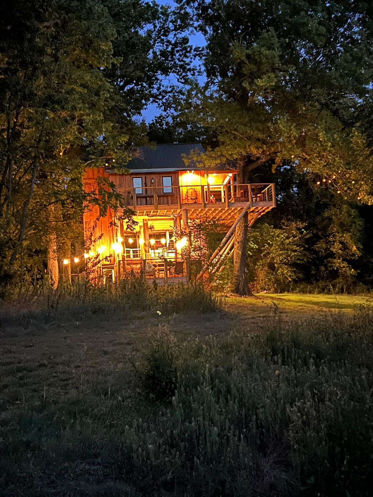 A custom-built cabin is illuminated at night, highlighting its multi-level structure nestled among trees. Warm lights reflect off the wood exterior, creating a welcoming ambiance. The outdoor spiral staircase and deck reveal a tranquil setting surrounded by nature.